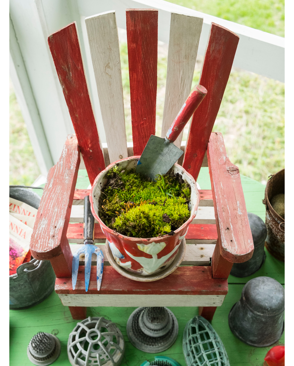 Un jardin secret de mousse nichée dans un pot de fleurs vintage prend place dans un fauteuil Adirondack miniature entourée d'une collection de pique-fleurs