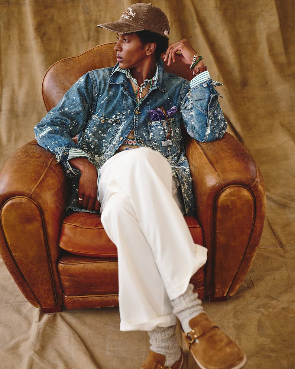 Homme assis, portant une veste en denim, une chemise à motifs, un pantalon blanc et une casquette.