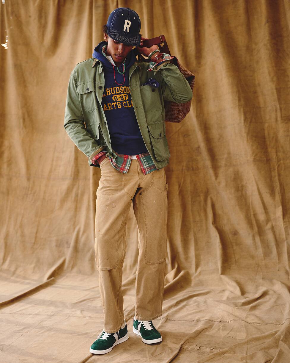 Un homme portant une veste utilitaire par-dessus un sweat à capuche et une chemise à carreaux, avec un pantalon beige, des baskets et une casquette.