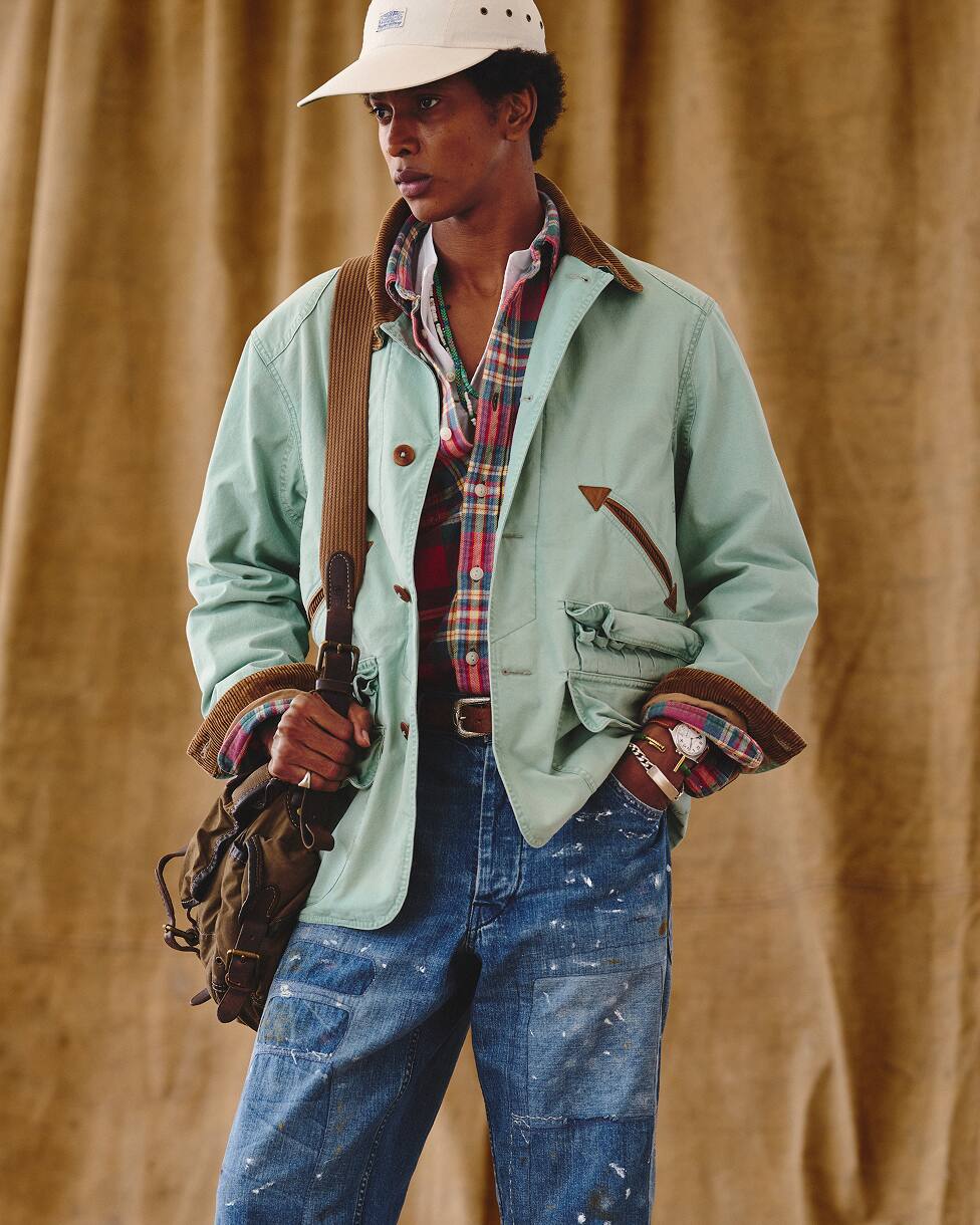 Un homme portant une veste verte sur une chemise à carreaux, un jean bleu, une casquette crème, avec un sac.