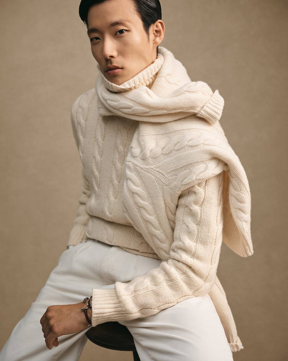 Homme assis portant un pull torsadé crème, une écharpe assortie et un pantalon blanc.