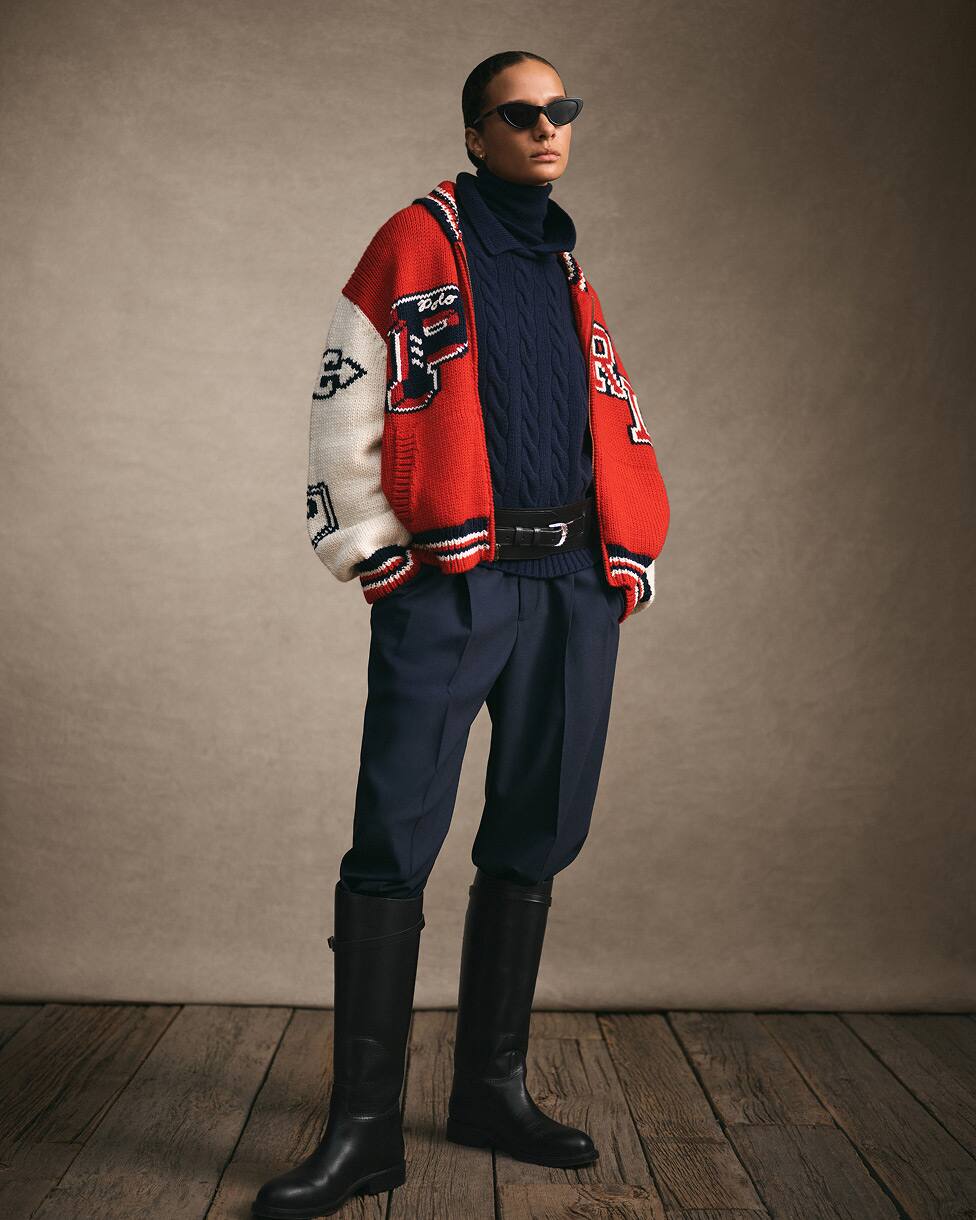 Femme debout portant une veste universitaire rouge et blanche, un pull et un pantalon marine et de hautes bottes noires.